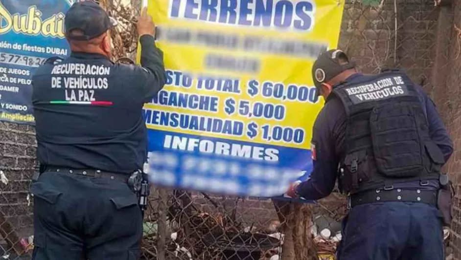Dos oficiales de recuperación de vehículos colocando un cartel informativo sobre terrenos en una valla.