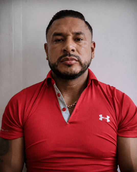 Hombre con barba y cabello corto, vistiendo una camiseta roja de cuello con detalles grises. Fondo blanco.