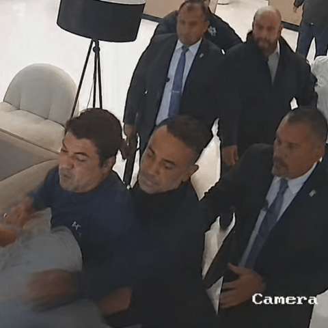 magen de videovigilancia que muestra a varias personas involucradas en una confrontación dentro de un inmueble. Se observa a un grupo de hombres sujetando a otra persona mientras varios individuos observan la escena en un pasillo interior.