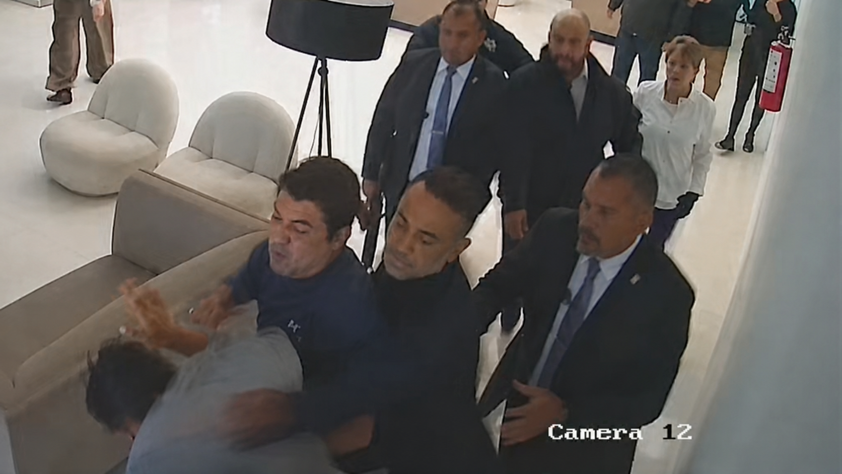 magen de videovigilancia que muestra a varias personas involucradas en una confrontación dentro de un inmueble. Se observa a un grupo de hombres sujetando a otra persona mientras varios individuos observan la escena en un pasillo interior.