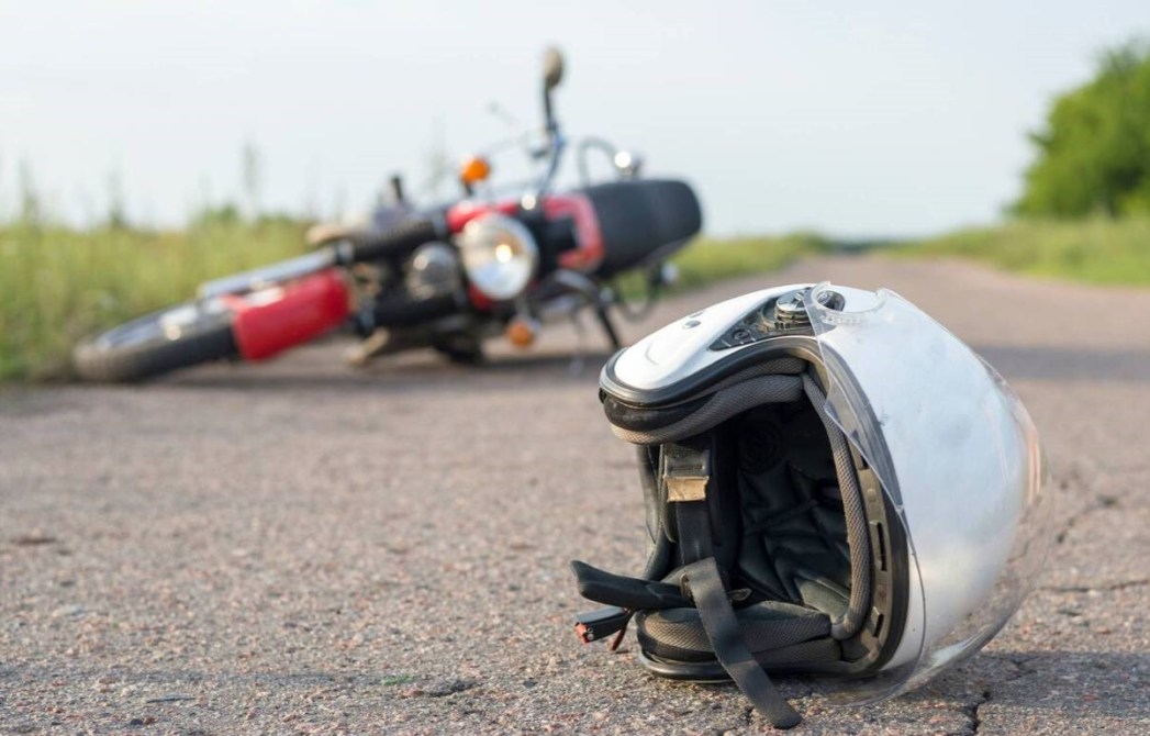 Un casco de moto en primer plano con una motocicleta caída al fondo en una carretera.