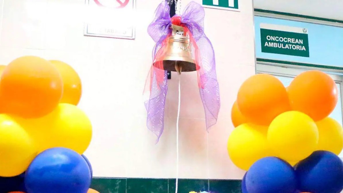 Campana dorada decorada con un moño morado en un ambiente hospitalario, flanqueada por globos de colores naranja, amarillo y azul.