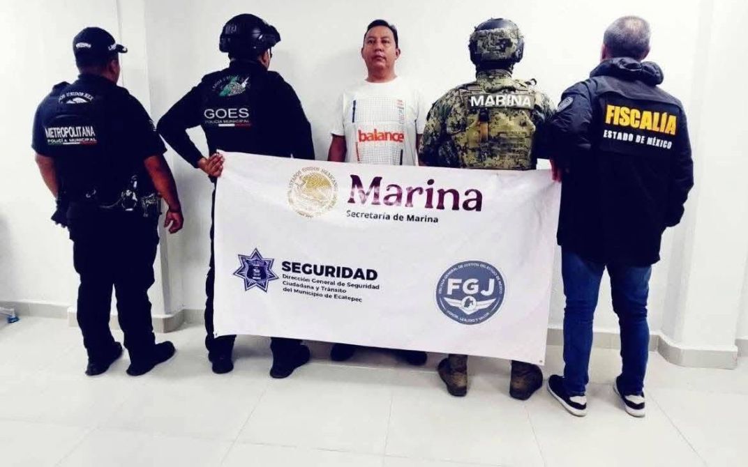 Detención de un sospechoso en presencia de autoridades de seguridad, con un cartel de la Secretaría de Marina y otras instituciones en un ambiente cerrado.